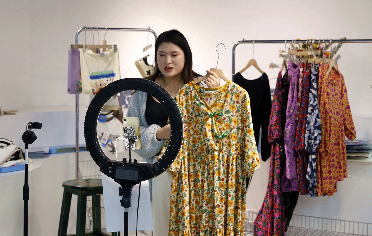 Presenter mengangkat gaun bermotif bunga di depan ring light dan rak baju